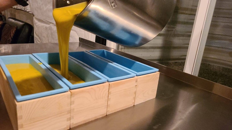 May include: A person pouring yellow liquid into five blue silicone molds, each sitting in a wooden box. The molds are lined up on a stainless steel surface.