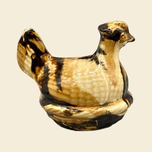 May include: A ceramic chicken figurine with brown and white glaze. The chicken is sitting on a nest and has a detailed head and beak.