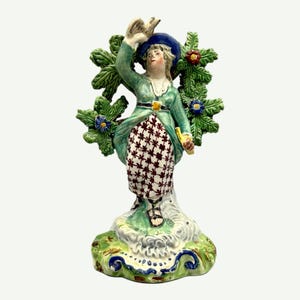 May include: A vintage porcelain figurine of a man in a green coat, blue hat, and patterned skirt, holding a bird. The figurine is surrounded by a green wreath with red and blue flowers, standing on a decorative base.