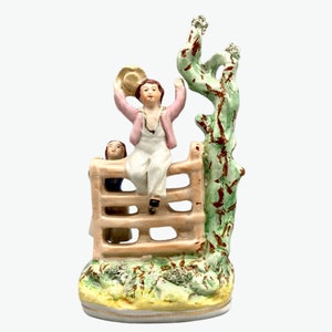 May include: A vintage ceramic figurine of two children sitting on a fence. The boy is wearing a pink jacket and a straw hat, while the girl is wearing a blue dress. The fence is painted brown and has a green tree behind it.