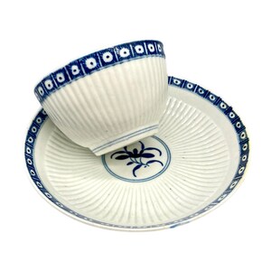 May include: A white teacup and saucer with blue and white floral designs. The cup and saucer have a fluted design and a blue border with white circles.