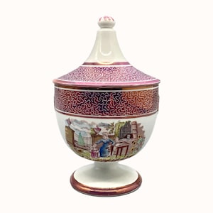 May include: A white ceramic sugar bowl with a pink and gold lid. The bowl features a floral design and a scene of a woman and a child in a garden. The bowl sits on a white pedestal.