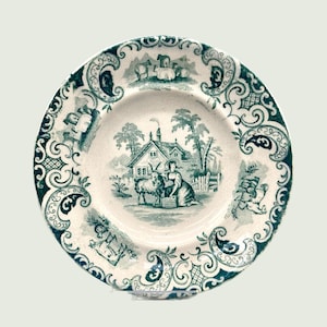 May include: A white ceramic plate with a green and white design. The plate features a scene of a woman milking a goat in front of a small cottage. The border of the plate is decorated with a repeating pattern of green swirls.