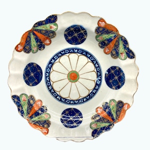 May include: A white ceramic plate with a scalloped edge and gold trim. The plate features a central floral design with orange and green accents, surrounded by blue and gold starburst patterns and fan-shaped designs.