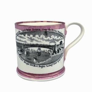 May include: A white ceramic mug with a pink rim and handle. The mug features a black and white illustration of a cast iron bridge over a river with ships and buildings. The text on the mug reads "View of the Cast Iron Bridge. Over the River Wear. 2.36 Feet Height 100 Feet Begun 24 Sep. 1793 Opened."