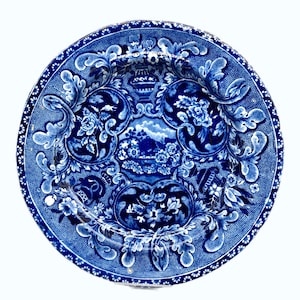 Peut inclure: Une assiette ronde en céramique bleue et blanche avec un motif floral et paysager détaillé. L'assiette présente une scène de paysage centrale entourée de motifs floraux ornés et d'une bordure pointillée. L'assiette mesure environ 25 cm de diamètre.
