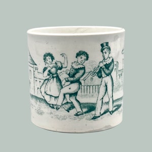 May include: A white ceramic mug with a green transferware design depicting three figures. A woman in a dress dances with a man, while another man plays a musical instrument. The scene is set against a backdrop of buildings.