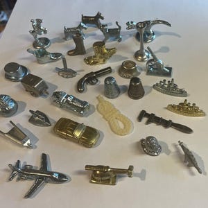 May include: A collection of Monopoly game tokens in various metallic finishes. The tokens include a car, airplane, dog, boot, thimble, cannon, and more. The tokens are arranged on a white surface.