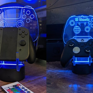 May include: Two illuminated gaming controller stands with a blue glow. The stand on the left features a Nintendo Switch controller and a remote control. The stand on the right features an Xbox controller and the text "Hi Lomein" on the top.