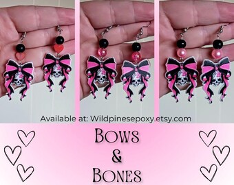 May include: Pink and black skull and bow earrings. The bows are pink with white polka dots and the skulls are black with white details. The earrings are available at Wildpinesepoxy.etsy.com. Bows & Bones.