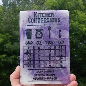 May include: A rectangular kitchen conversion chart made of purple and white marbled resin. The surface features black text and icons for cups, ounces, tablespoons, and teaspoons, including a conversion table and volume measurements in cups, pints, and gallons.
