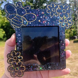 May include: A decorative resin photo frame with a bee and flower design. The frame is black with gold accents and glitter. The bee and flowers are outlined in gold. The frame is held in a hand.