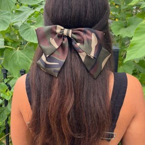May include: A large bow hair clip in a camouflage pattern is secured in long, dark brown hair. The bow features shades of green, brown, and tan. The background includes sunflowers and green foliage, suggesting an outdoor setting. The hair accessory adds a stylish touch.