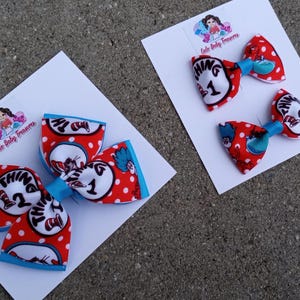 May include: Two red and white bow hair clips with blue accents. The bows feature a white background with red polka dots and a graphic of the character Thing 1 and Thing 2 from the book "The Cat in the Hat".