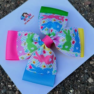 May include: A large, colorful hair bow with an Easter theme. The bow features a pattern of bunnies and the word "Easter" on a white background. The bow has pink, green, blue, and yellow accents. The center of the bow is tied with a pink ribbon.