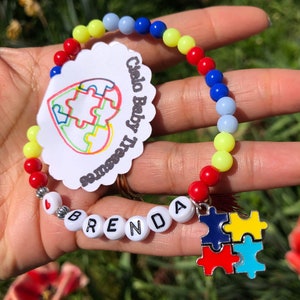 May include: A colorful beaded bracelet with the name "BRENDA" spelled out in white letter beads. The bracelet features a puzzle piece charm and a tag that says "Cielo Baby Treasures". The beads are red, yellow, blue, and light blue.