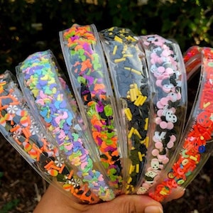 May include: A collection of clear plastic headbands filled with colorful Halloween-themed confetti. The confetti includes bats, pumpkins, candy corn, and other festive shapes in orange, black, pink, and yellow. The headbands are held in a hand.