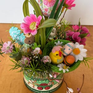 Può includere: Una lattina verde con un motivo floreale e un'immagine di un carro pieno di mele. La lattina è piena di una varietà di decorazioni pasquali, tra cui un nido con un pulcino giallo e uova a strisce blu e bianche, fiori rosa, erba verde e un uovo con motivo rosa e bianco.