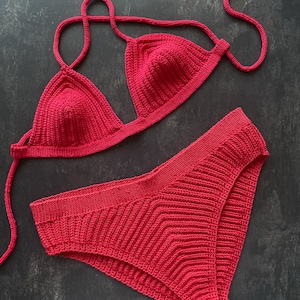 Könnte beinhalten: Ein leuchtend rotes Häkel-Bikini-Set. Das Oberteil hat Triangel-Cups mit verstellbaren Trägern, und das Unterteil ist hoch tailliert mit einem strukturierten Muster. Das Set ist aus weichem, gestricktem Material gefertigt, ideal für Bademode.