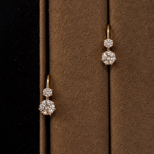 May include: A pair of gold drop earrings, each featuring two round, brilliant-cut clear stones set in a prong setting. The smaller stone is positioned above the larger stone, with both suspended from a curved gold wire hook mechanism.