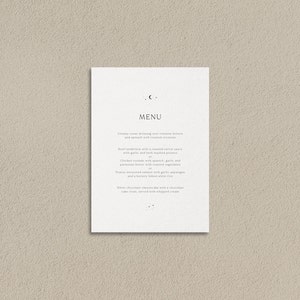 May include: A white menu card with a simple design featuring a crescent moon symbol and a list of food items. The menu includes creamy Caesar dressing, beef tenderloin, chicken roulade, peanut encrusted salmon, and white chocolate cheesecake.