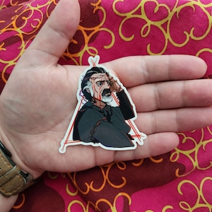 May include: A sticker featuring a detailed illustration of a man with a beard and a crown of thorns. He is wearing a dark grey shirt and has a red triangle around him. The sticker is on a hand.