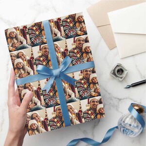 K&ouml;nnte beinhalten: Ein Blatt Geschenkpapier mit einem sich wiederholenden Muster einer dreik&ouml;pfigen Familie, die f&uuml;r ein Weihnachtsfoto posiert. Die Familie tr&auml;gt festliche Kleidung und l&auml;chelt. Das Geschenkpapier ist mit einem blauen Band gebunden.