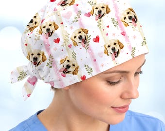May include: A white scrub cap featuring a repeating pattern of golden retriever dog faces, pink watercolor stripes, small red hearts, and green leafy sprigs. The cap includes a tie-back closure at the nape of the neck for an adjustable fit.