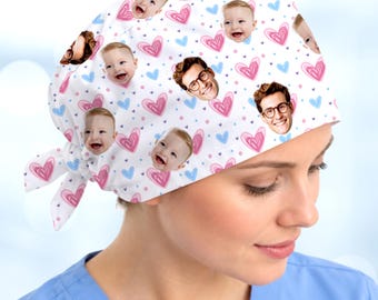 May include: A white scrub cap featuring a repeating pattern of pink and blue hand-drawn hearts, small blue dots, and photographic portraits of a smiling baby and a man wearing glasses. The cap includes a tie-back closure at the nape.