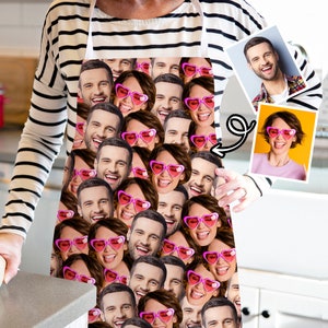 May include: A white apron with a pattern of multiple faces wearing pink heart-shaped sunglasses. The faces are all different and some are smiling, while others are sticking their tongues out.