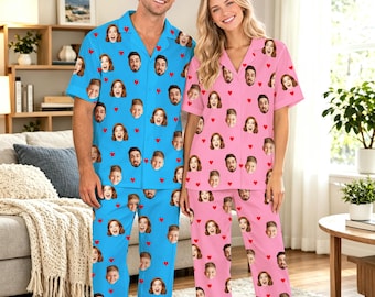May include: A set of matching pajamas featuring a blue button-down shirt and pants for one person, and a pink button-down shirt and pants for another. Both sets are covered in a repeating pattern of small red hearts and a person's face.