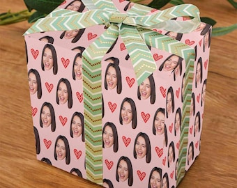 May include: A pink gift box with a repeating pattern of a woman's face winking and red hearts. The box is tied with a green and gold chevron patterned ribbon, creating a festive and personalized presentation.