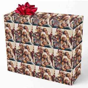K&ouml;nnte beinhalten: Ein Weihnachtsgeschenkpapier mit einem sich wiederholenden Muster einer dreik&ouml;pfigen Familie, einem Mann, einer Frau und einem Kind, die Weihnachtsm&uuml;tzen tragen und l&auml;cheln. Die Familie sitzt vor einem beleuchteten Weihnachtsbaum.