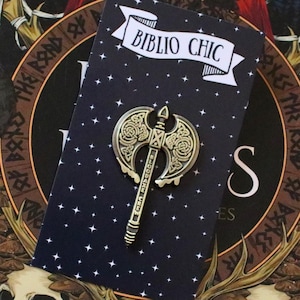 May include: A gold-colored axe pin with intricate Celtic knotwork, displayed on a navy blue card with the text "BIBLIO CHIC". The pin is set against a book cover featuring a woman in a red hood and skull imagery.