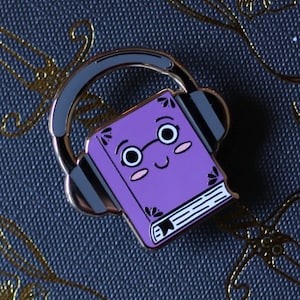 Peut inclure: Livre violet avec un visage souriant portant un casque. Le livre a un contour doré et une tranche blanche avec du texte noir.