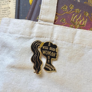 May include: A black and gold enamel pin featuring a woman's silhouette with long hair. The pin reads "A Well Read Woman Is A Dangerous Creature" in white text. The pin is on a white tote bag.