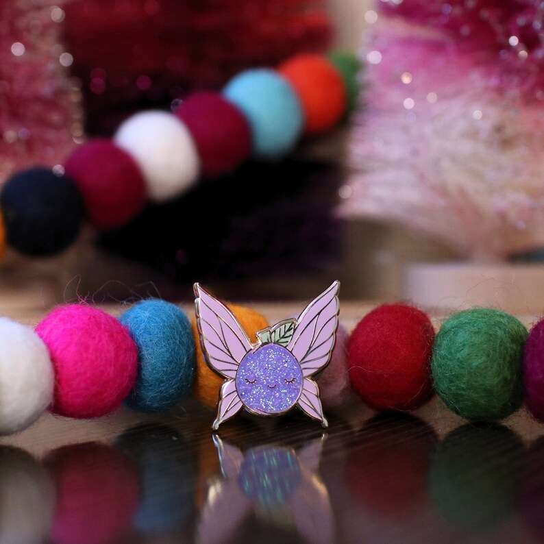 May include: Purple glitter enamel pin with a smiling face and wings. The pin is shaped like a fairy and has a small green leaf on top of its head.