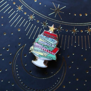 May include: A gold-tone enamel pin shaped like a Christmas tree made of stacked books. The books are decorated with red, green, blue, and white patterns. The top book has a red cover with a gold star on top. The pin says "Merry Christmas" on one of the books.
