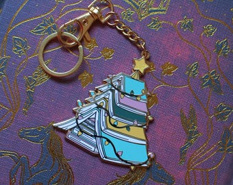 May include: A gold-tone keychain shaped like a Christmas tree made of stacked books with a star on top. The books are decorated with colorful lights and have a blue, green, pink, and yellow color scheme.