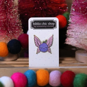 May include: A purple glitter fairy with pink wings and a gold outline. The fairy is smiling with closed eyes. The pin is on a white card with the text "biblio chic shop by sincerely sirena".