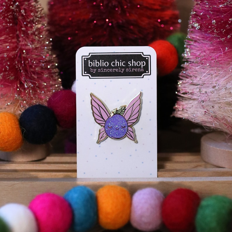 May include: A purple glitter fairy with pink wings and a gold outline. The fairy is smiling with closed eyes. The pin is on a white card with the text "biblio chic shop by sincerely sirena".