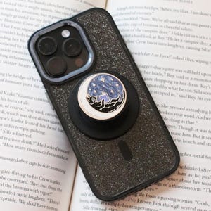 May include: A black glittery smartphone case with a circular phone grip attached to the back. The grip features a circular illustration of a black cat lying on a crescent moon against a blue starry night sky, while reading a small book.
