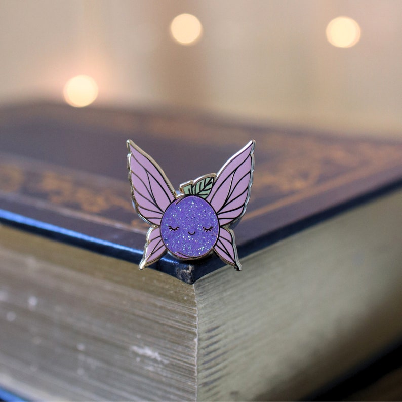 May include: A purple glitter fairy pin with pink wings and a smiling face. The pin is sitting on a closed book.
