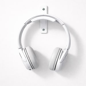 May include: White over-ear headphones suspended from a white wall-mounted holder. The headphones feature a padded headband and earcups. The holder is secured to the wall with screws, providing a clean, minimalist aesthetic.