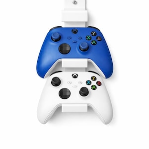 May include: A white wall mount holding two game controllers. The top controller is blue, and the bottom controller is white. Both controllers have black buttons and coloured action buttons.