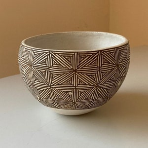 Puede incluir: Un cuenco de cerámica con un patrón geométrico. El cuenco es blanco por dentro y tiene un diseño triangular marrón y blanco en el exterior. El cuenco está sobre una superficie blanca.
