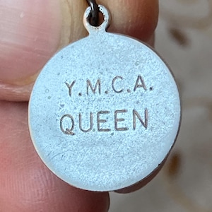 May include: A small, round, silver-toned charm with the text "Y.M.C.A. QUEEN" engraved on its surface. The charm has a small loop at the top for attaching to a chain or bracelet. The charm is held between fingers.