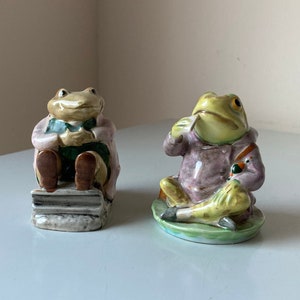 Könnte beinhalten: Zwei Keramik-Froschfiguren, eine sitzt auf einem Stapel Bücher und die andere auf einem Seerosenblatt. Beide Frösche tragen lila Jacken und haben grüne Augen.