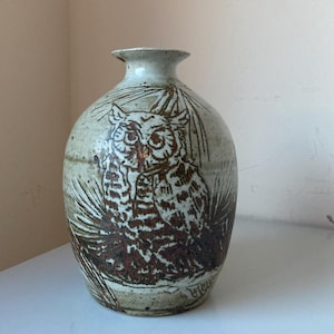May include: A ceramic vase with a rounded body and narrow neck. The vase features a brown owl illustration with detailed feathers and a branch design. The color palette includes shades of brown, beige, and cream.