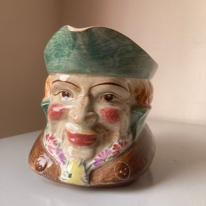 Puede incluir: Una jarra de cerámica con forma de cabeza de hombre con un sombrero verde, una chaqueta marrón y una bufanda floral. El hombre tiene una cara sonriente con mejillas sonrosadas.